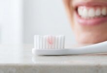 A macro photo showing a toothbrush with a faint red tint on its bristles, indicating an early sign of gum disease, with a person's healthy mouth blurred in the background.