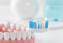 A close-up, photorealistic image of healthy, pink gums and clean white teeth. A soft-bristled toothbrush is gently placed near the gum line. In the blurred background, dental floss and a mouthwash bottle are visible, symbolizing comprehensive oral hygiene.