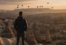 A person stands on a viewpoint overlooking the unique 'fairy chimney' rock formations of Cappadocia, Turkey, at sunrise. Colorful hot air balloons are visible in the distant sky. The scene evokes a sense of travel, adventure, and well-being.