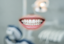 A close-up shot of a human mouth with a radiant, perfectly white, and symmetrical smile. The teeth are pristine and flawless, set against healthy, pink gums. The background is a soft, out-of-focus blur of a modern dental clinic, suggesting precision and advanced care. No text or identifying features are visible.