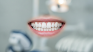 A close-up shot of a human mouth with a radiant, perfectly white, and symmetrical smile. The teeth are pristine and flawless, set against healthy, pink gums. The background is a soft, out-of-focus blur of a modern dental clinic, suggesting precision and advanced care. No text or identifying features are visible.