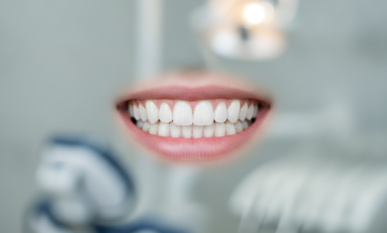 A close-up shot of a human mouth with a radiant, perfectly white, and symmetrical smile. The teeth are pristine and flawless, set against healthy, pink gums. The background is a soft, out-of-focus blur of a modern dental clinic, suggesting precision and advanced care. No text or identifying features are visible.