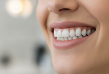 Close-up of a person smiling broadly, showing a full set of healthy, natural-looking, perfectly aligned teeth. The background is a soft, out-of-focus blur of a clean, modern dental office, conveying professionalism and successful dental restoration.